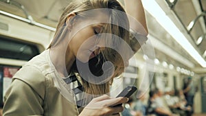 Portrait of a smiling lovely girl typing message on mobile phone in subway train, student blonde woman with