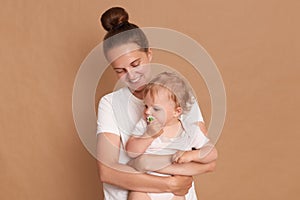 Portrait of smiling loving gentle mother hugging baby daughter  over brown background, mommy looking at kid with smile,