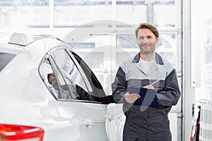 Portrait of smiling male automobile mechanic holding clipboard while standing by car in repair shop