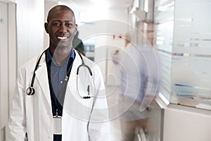 Portrait Of Smiling Male Doctor Wearing White Coat With Stethoscope In Busy Hospital Corridor