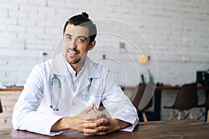 Portrait of smiling male doctor wearing white uniform with stethoscope sitting at desk in hospital office, friendly