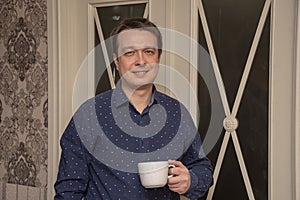 Portrait of a smiling man 30-35 years old with a mug in his hands on the background of home interior. Maybe he`s an actor or TV ho