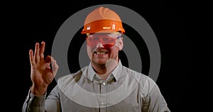 Portrait of a smiling man on a black background in the studio, wearing an construction helmet, protective glasses and a