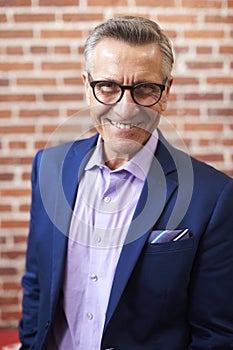 Portrait Of Smiling Mature Businessman Standing Against Brick Wall In Modern Office