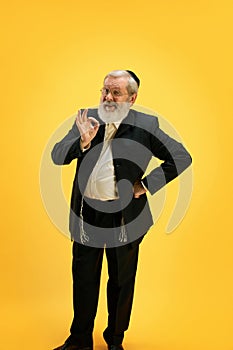 Portrait of smiling mature man in suit and black kippah making positive hand gesture against sunny yellow background