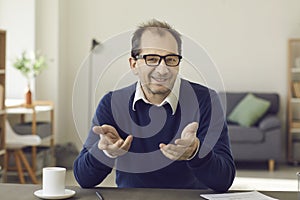 Portrait of smiling mature man at table with gesturing hand looking at camera