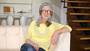 Portrait of smiling mature woman in casual wear and glasses sitting on the white sofa