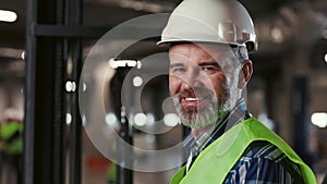 Portrait of Smiling Mature Worker in Safety Vest and Hard Hat. Forklift Driver Looking at Camera. Warehouse with Shelves