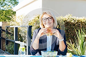 Portrait of smiling middle-aged business woman eating healthy burger during lunch at the park outdoor cafe during her