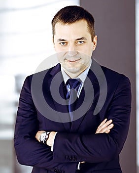 Portrait of a smiling middle aged businessman in a dark blue suit, standing with crossed arms at meeting in an office