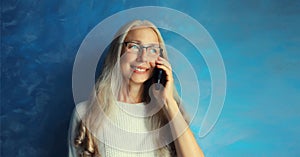 Portrait of smiling middle-aged gray-haired woman calling on mobile phone in eyeglasses on blue background