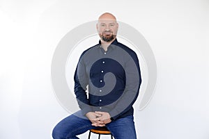 Portrait of smiling middle-aged man wear blue shirt, jeans sitting on stool, interlacing fingers on white background.