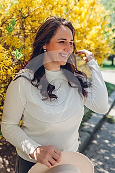 Portrait of smiling middle-aged woman walking in blooming spring garden. Happy stylish lady holding hat outdoors