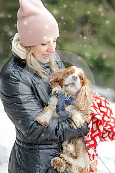 Portrait of smiling middle-aged woman wearing black jacket, holding looking at dog spaniel in forest in snowy winter.