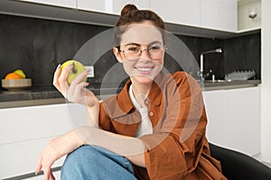Portrait of smiling, modern woman in glasses, sitting at home and eating green apple, having a lunch break, biting fruit