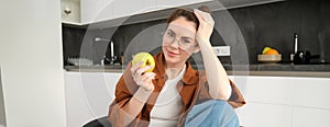 Portrait of smiling, modern woman in glasses, sitting at home and eating green apple, having a lunch break, biting fruit