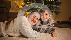 Portrait of smiling mom with son embracing and lying on carpet under Christmas tree in living room. Pure emotions of