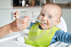 Portrait Of Smiling 8 month Old Baby Boy At Home In High Chair Being Fed Solid Food By Mother With Spoon