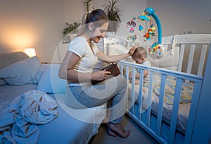 Portrait of young smiling mother in pajamas sitting on bed and reading book to her baby son before going to sleep