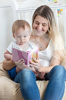 Portrait of smiling young mother sitting on beanbag with her baby boy and reading him book