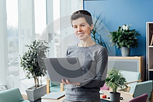 Portrait of Smiling Neutral gender middle aged woman in casual outfit working on laptop while standing in modern office