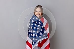 Patriotic little girl standing wrapped in american flag, looking at camera, relocating to America.