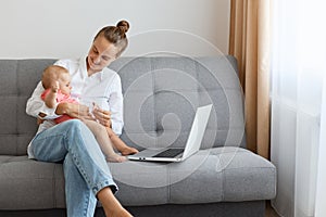 Portrait of smiling positive young mother with baby, woman with bun hairstyle wearing white shirt and jeans playing with toddler