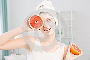Portrait of smiling pretty girl with white towel on head holding halves of grapefruits near face closing one eye