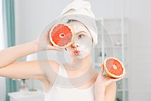 Portrait of smiling pretty girl with white towel on head holding halves of grapefruits near face closing one eye