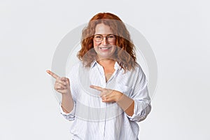 Portrait of smiling pretty middle-aged woman with red hair, wearing glasses, showing way, helping with choice