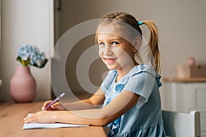 Portrait of smiling pretty primary girl child writing doing homework sitting at home table by window looking at camera.