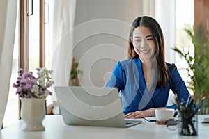 Portrait of smiling pretty young asian business woman sitting on workplace and using laptop computer.