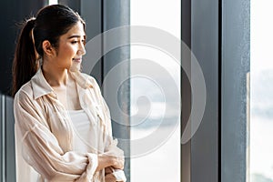 Portrait of smiling pretty young business woman standing on workplace office and looking out to window.Smile and Thinking