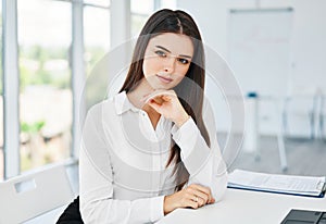 Portrait of smiling pretty young businesswoman sitting on workplace at the table in modern office and looking at camera