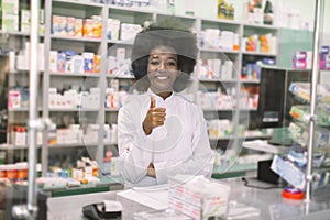 Portrait of smiling professional African female pharmacist in modern drugstore, standing behind the glass at the counter