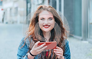 Portrait of smiling red curled long hair caucasian teen girl walking on the street and video chatting using the modern smartphone