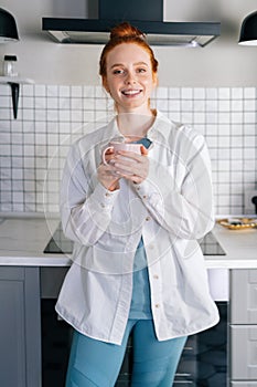 Portrait of smiling redhead woman holding cup of hot beverage in hands on morning at kitchen.