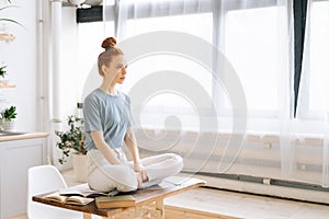 Portrait of smiling redhead young woman meditating while sits on desk at home office