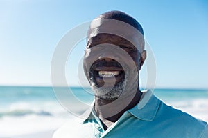 Portrait of smiling retired african american senior bald man at beach against blue sky on sunny day