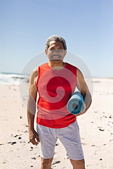 Portrait of smiling retired biracial man holding rolled yoga mat at beach against blue sky