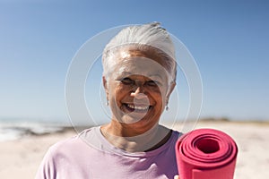 Portrait of smiling retired biracial senior woman holding exercise mat with short hair at beach