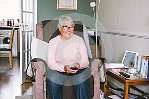 Portrait of smiling retired elderly woman in comfortable armchair with TV remote control and watching television channel
