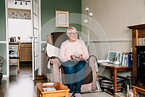 Portrait of smiling retired elderly woman in comfortable armchair with TV remote control and watching television channel