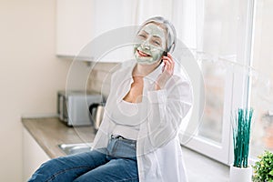 Portrait of smiling retired gray haired woman with green mud antiwrinkle facial mask sitting on kitchen countertop and photo