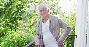 Portrait of smiling retired senior man wearing cardigan sweater leaning on railing in balcony