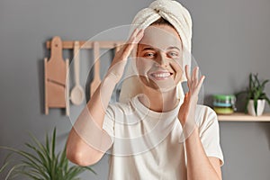 Portrait of smiling satisfied Caucasian woman with cosmetic mask for face skin posing in kitchen enjoying spa beauty procedures at