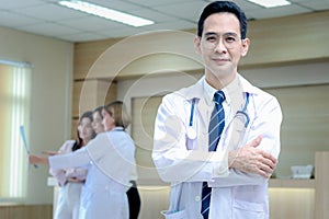 Portrait of smiling senior Asian male doctor in workwear with stethoscope, standing with arms crossed in clinic hospital, looks at