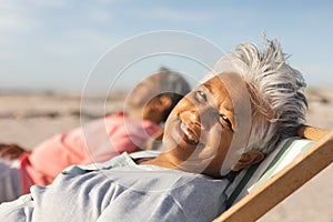 Portrait of smiling senior biracial woman relaxing on chair with man at beach during sunny day