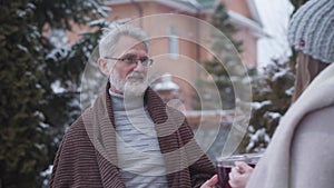 Portrait of smiling senior Caucasian man in eyeglasses talking to young woman bringing tea cups. Happy couple with age