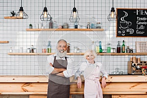 portrait of smiling senior coffee shop owners in aprons standing at counter